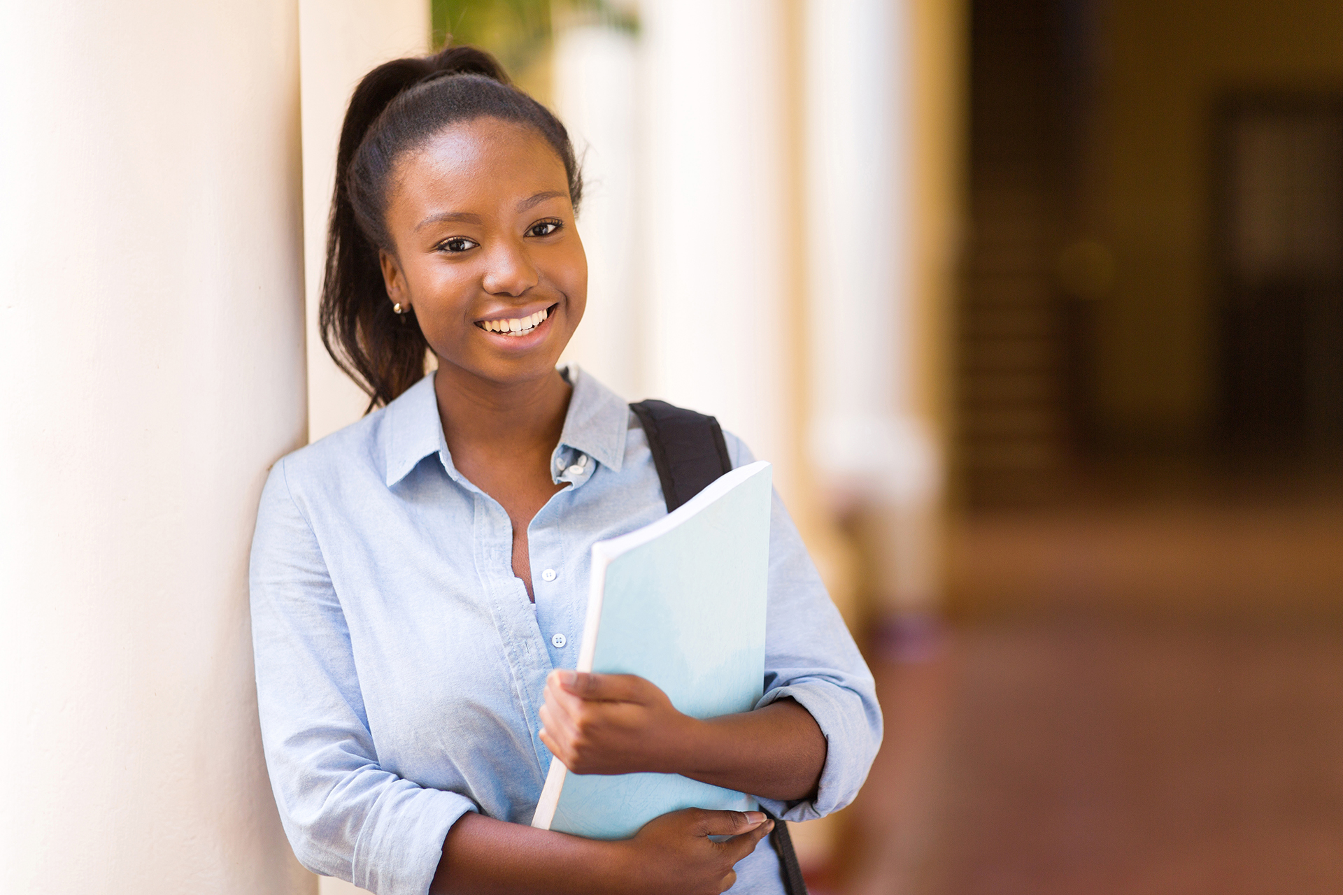 Photograph showing a smiling female African American college student leaning against a wall holding a folder.